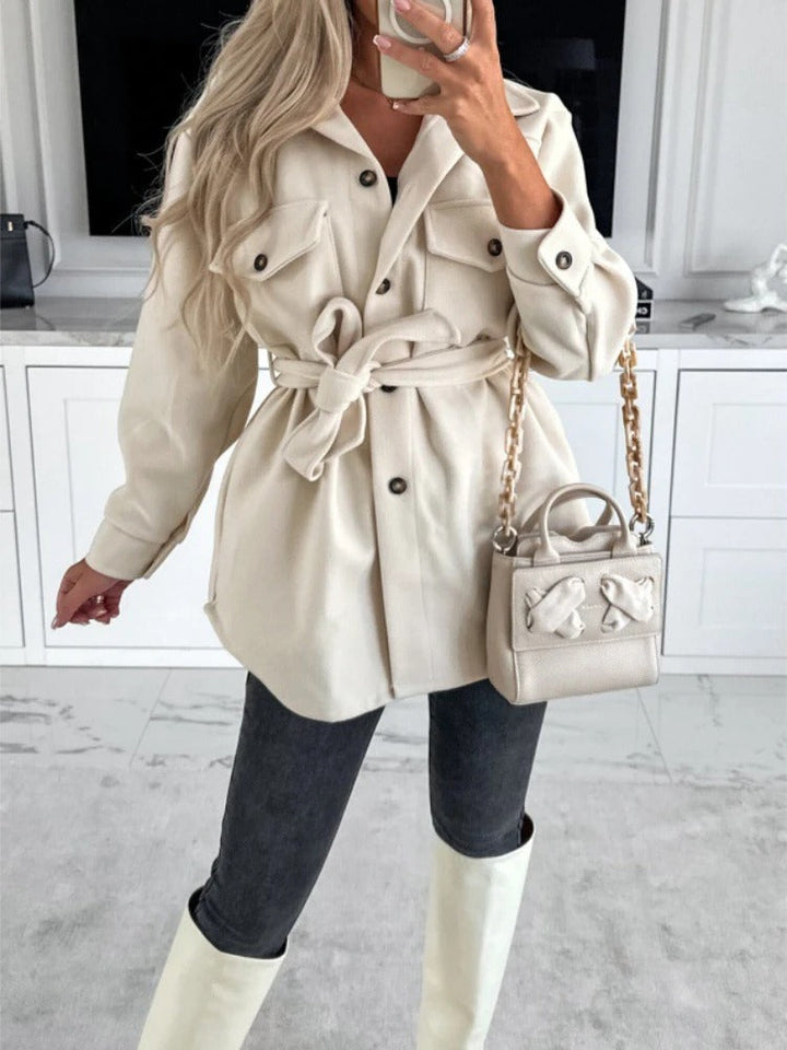 Frau in cremefarbener Jacke mit Gürtel, graue Jeans, weiße Stiefel, beige Handtasche.