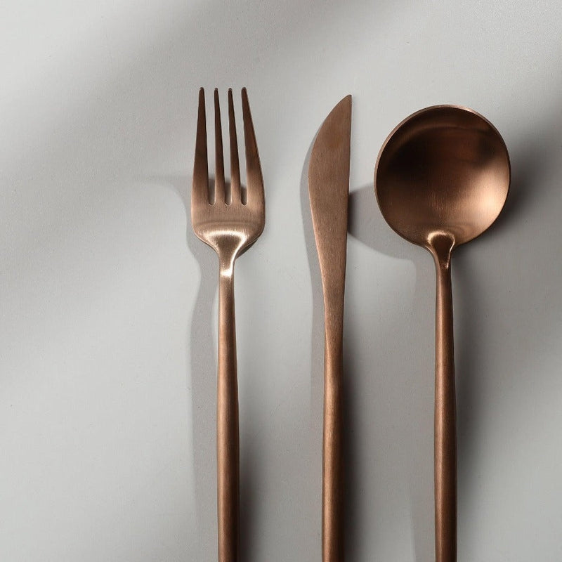 Minimalistisches Besteckset aus Kupfer, bestehend aus Gabel, Messer und Löffel auf hellem Hintergrund.