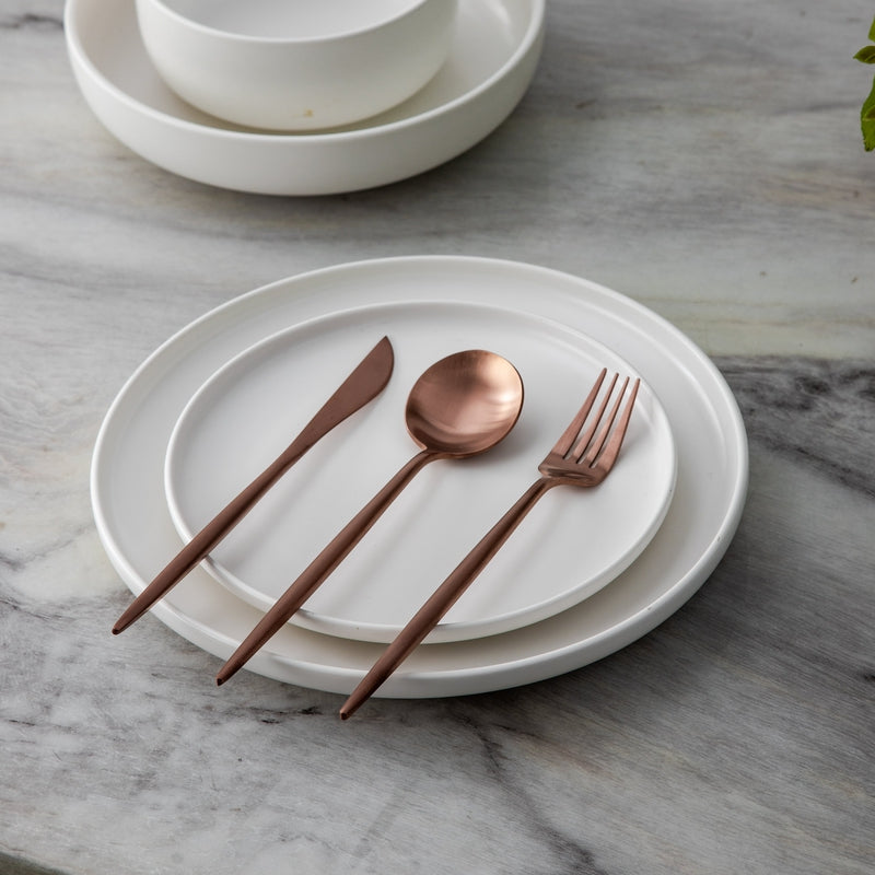 Weißes Keramikgeschirr mit roségoldenem Besteck auf Marmortisch.