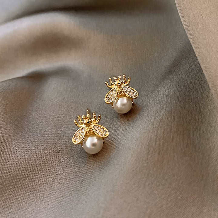 Goldene Bienen-Ohrringe mit Perlen und Strasssteinen auf grauem Stoff.