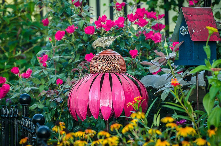 Rosa Metall-Vogelfutterspender im Garten mit Blumen und Vogel, dekorativer Futterspender.