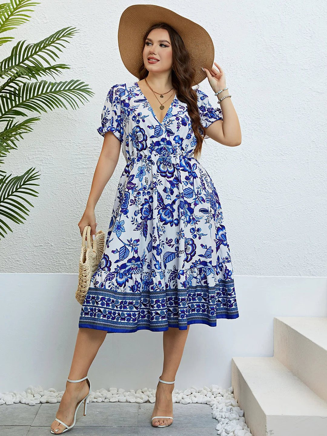 Frau im blauen Blumen-Sommerkleid mit Strohhut, Sandalen und Tasche vor weißer Wand.