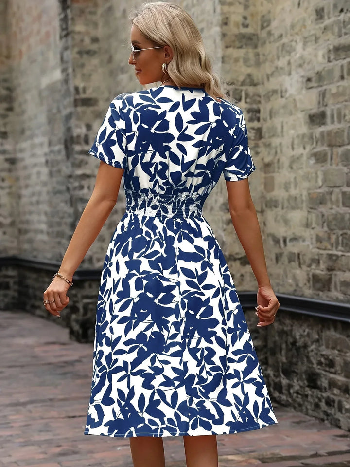 Frau in blau-weißem Sommerkleid mit Blattmuster, steht vor einer Backsteinmauer.