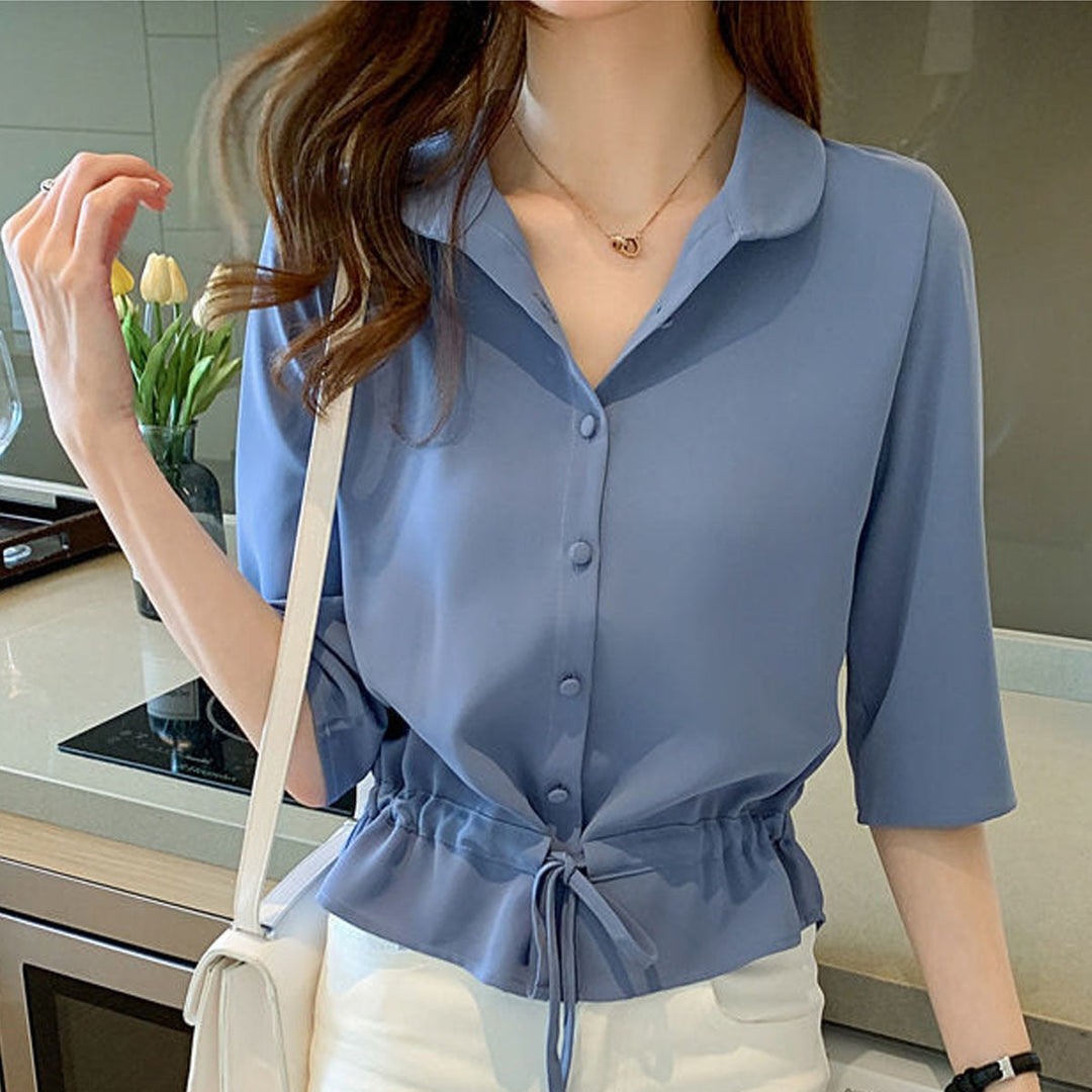 Frau in blauer Bluse mit Kordelzug, weiße Hose, moderne Küche, elegantes Outfit.