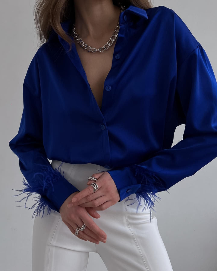 Frau in blauer Satinbluse mit Federn, weiße Hose, silberne Kette, modisches Outfit.