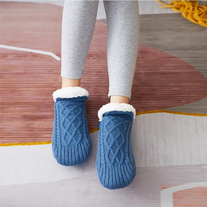 Blaue gestrickte Hausschuhe mit weißem Futter, getragen von Person in grauen Leggings.