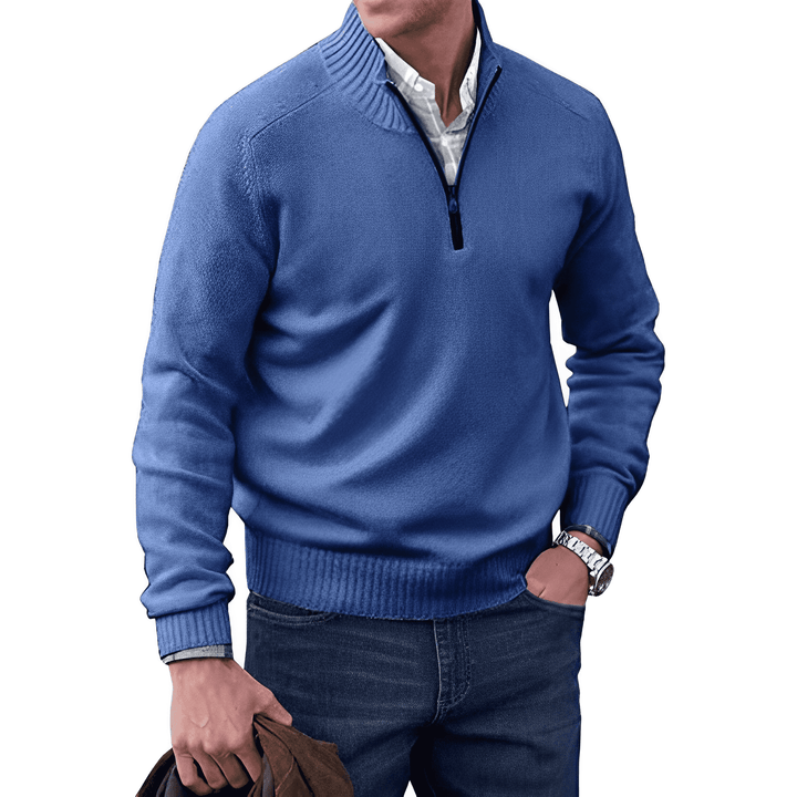 Mann in blauem Pullover mit Reißverschluss, weißem Hemd und Jeans, hält braune Jacke.
