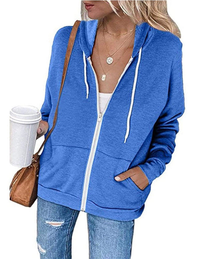 Frau in blauer Kapuzenjacke mit Reißverschluss, hält Kaffeebecher, trägt Jeans und braune Tasche.