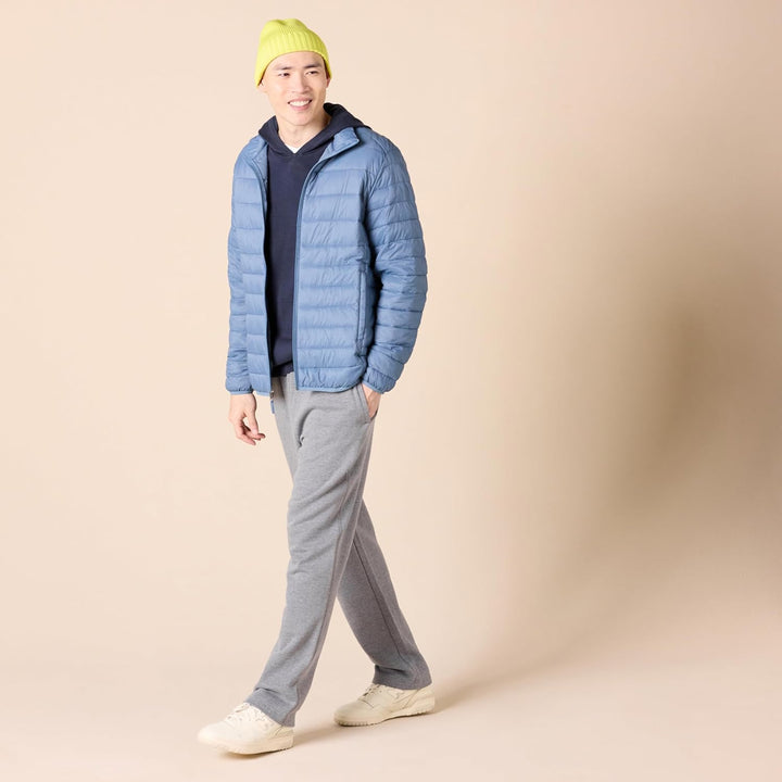Mann in blauer Steppjacke, grauer Jogginghose und gelber Mütze, lächelnd, beige Hintergrund.