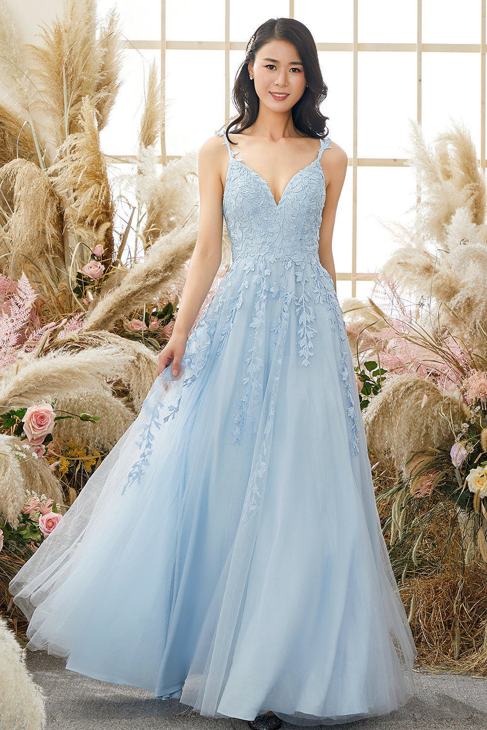 Frau im hellblauen Abendkleid mit Spitzen, vor Fenster und Pampasgras, elegantes Design.