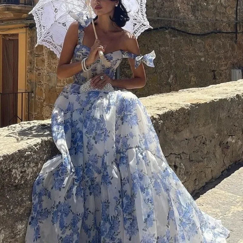 Frau in blau-weißem Blumenkleid mit Spitze, hält weißen Sonnenschirm, sitzt auf Steinmauer.