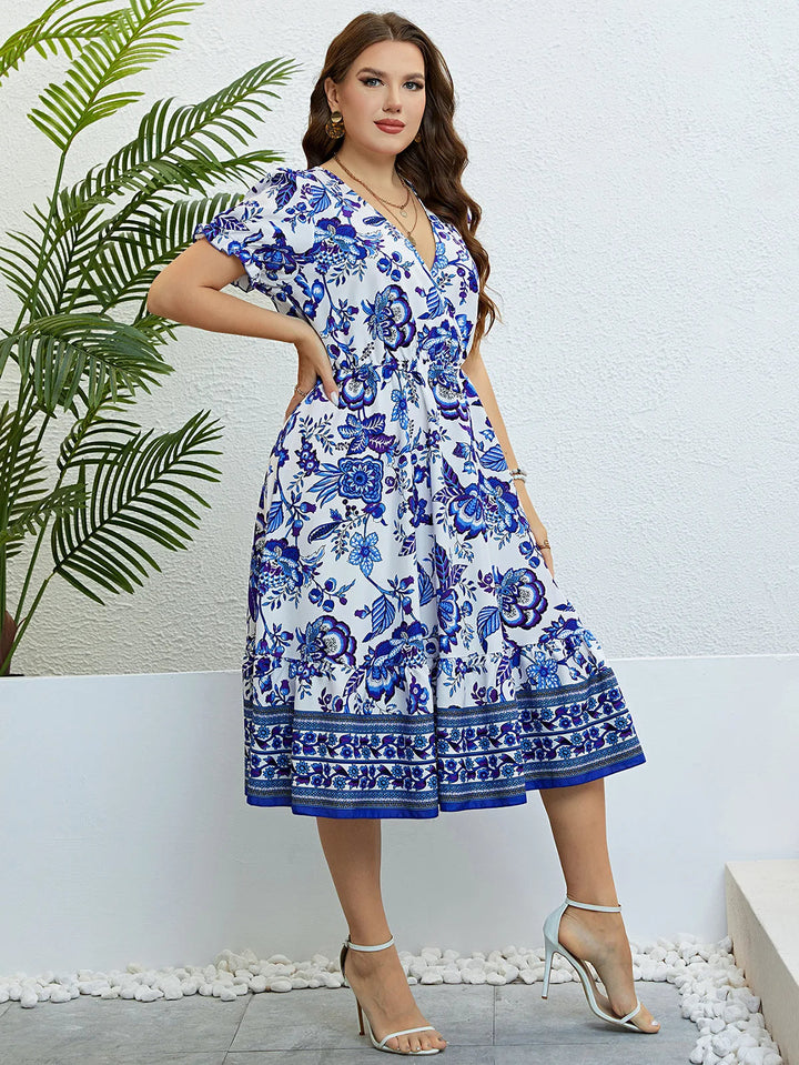 Frau in weißem, geblümtem Sommerkleid mit blauen Mustern, steht vor weißer Wand.