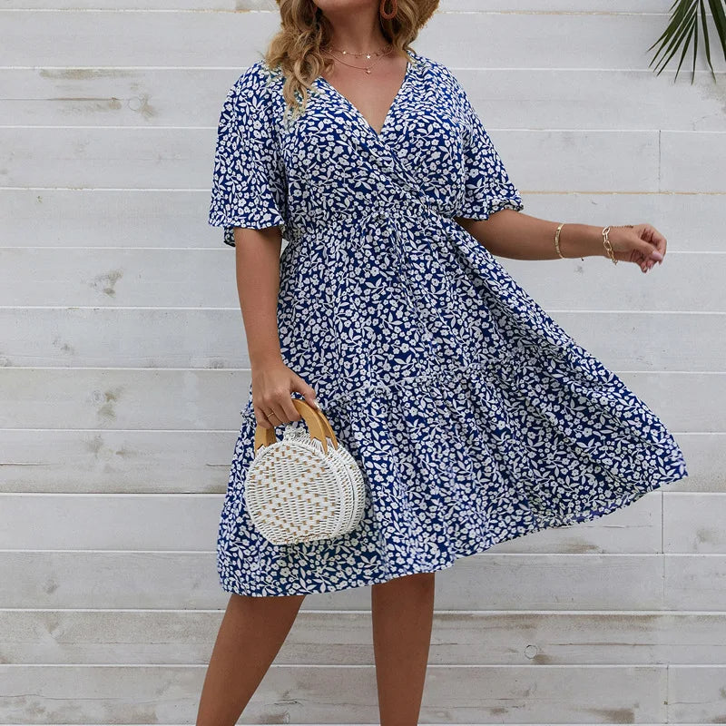 Frau im blauen Sommerkleid mit Blumenmuster, hält runde Strohtasche, vor weißer Holzwand.