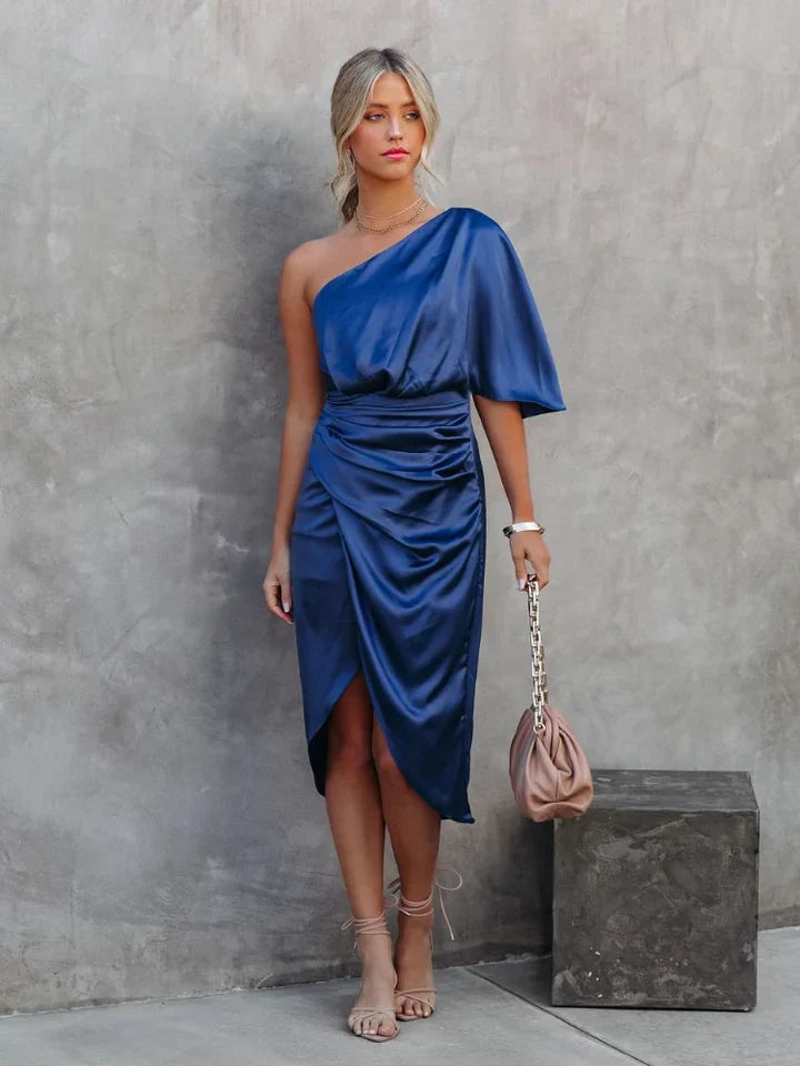 Frau in blauem, einärmeligem Satin-Kleid, hält beige Tasche, steht vor grauer Wand.