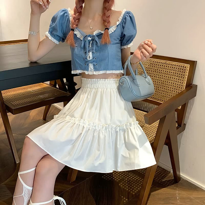 Frau in blauem Crop-Top und weißem Rock, hält eine kleine blaue Handtasche, sitzt auf Holzstuhl.