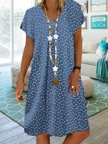 Frau in blauem Sommerkleid mit Herzmuster und Holzperlenkette im Wohnzimmer.