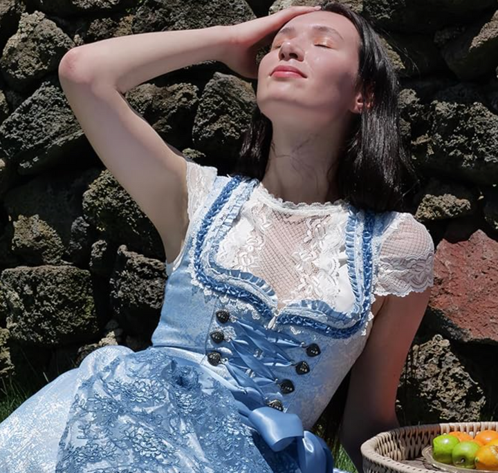 Frau in blauem Dirndl mit Spitzenbluse, sitzt vor Steinmauer, hält Gesicht in die Sonne.