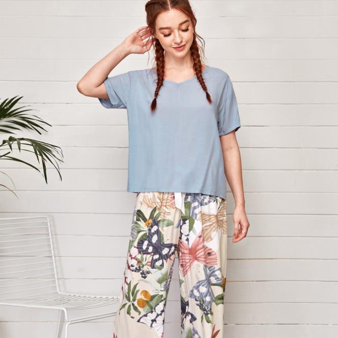 Frau in blauem T-Shirt und bunter Blumenhose vor weißer Wand, lässige Sommermode.