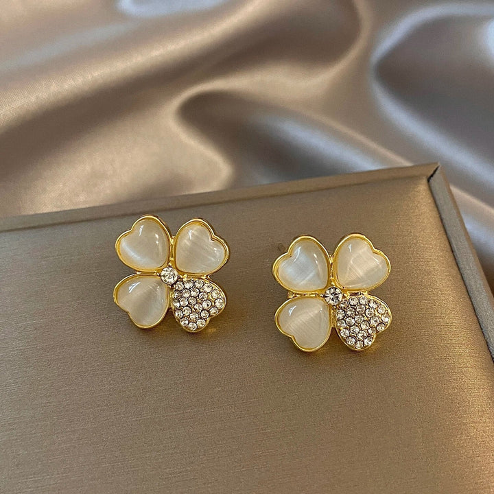 Goldene Ohrringe mit Herzblüten-Design, Perlmutt und Strasssteinen auf Seidenhintergrund.