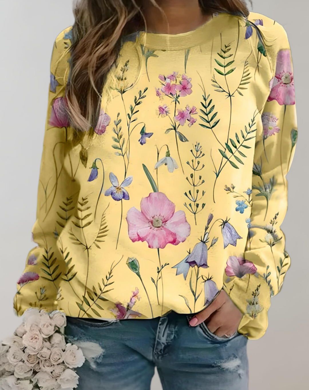 Frau in gelbem Blumenpullover, Jeans, hält weißen Blumenstrauß.