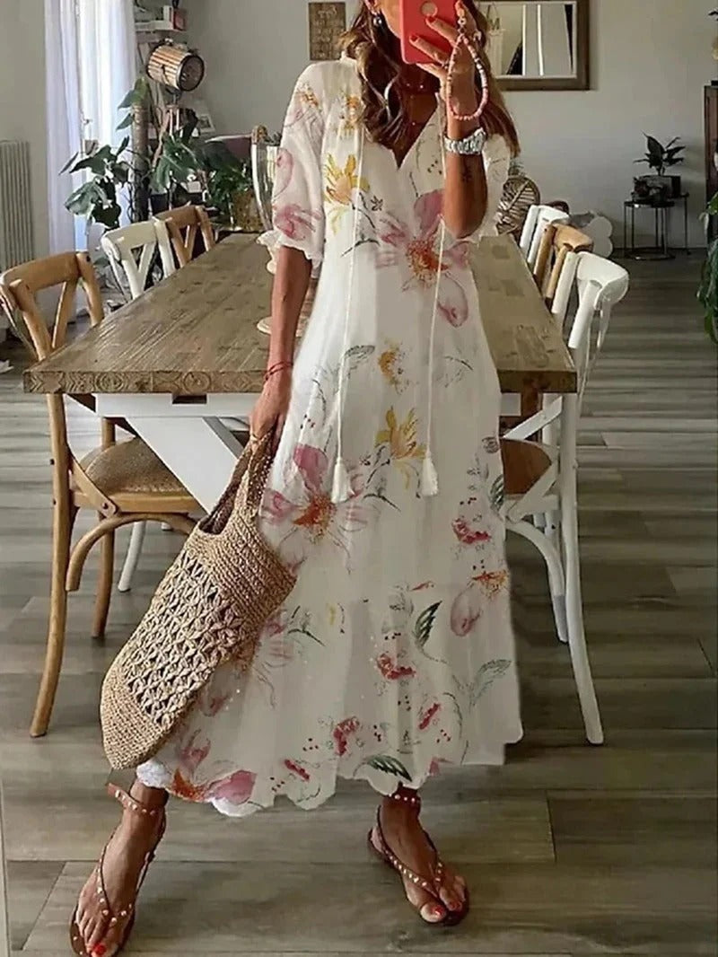 Frau in weißem, geblümtem Maxikleid mit Strohtasche, steht in modernem Esszimmer.