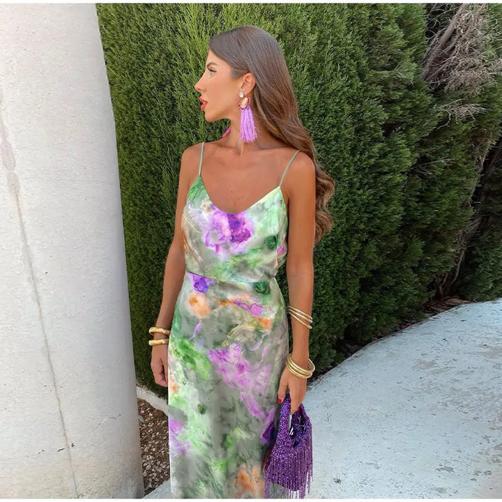 Frau in buntem Sommerkleid mit lila Quastenohrringen und Tasche, vor grüner Hecke.