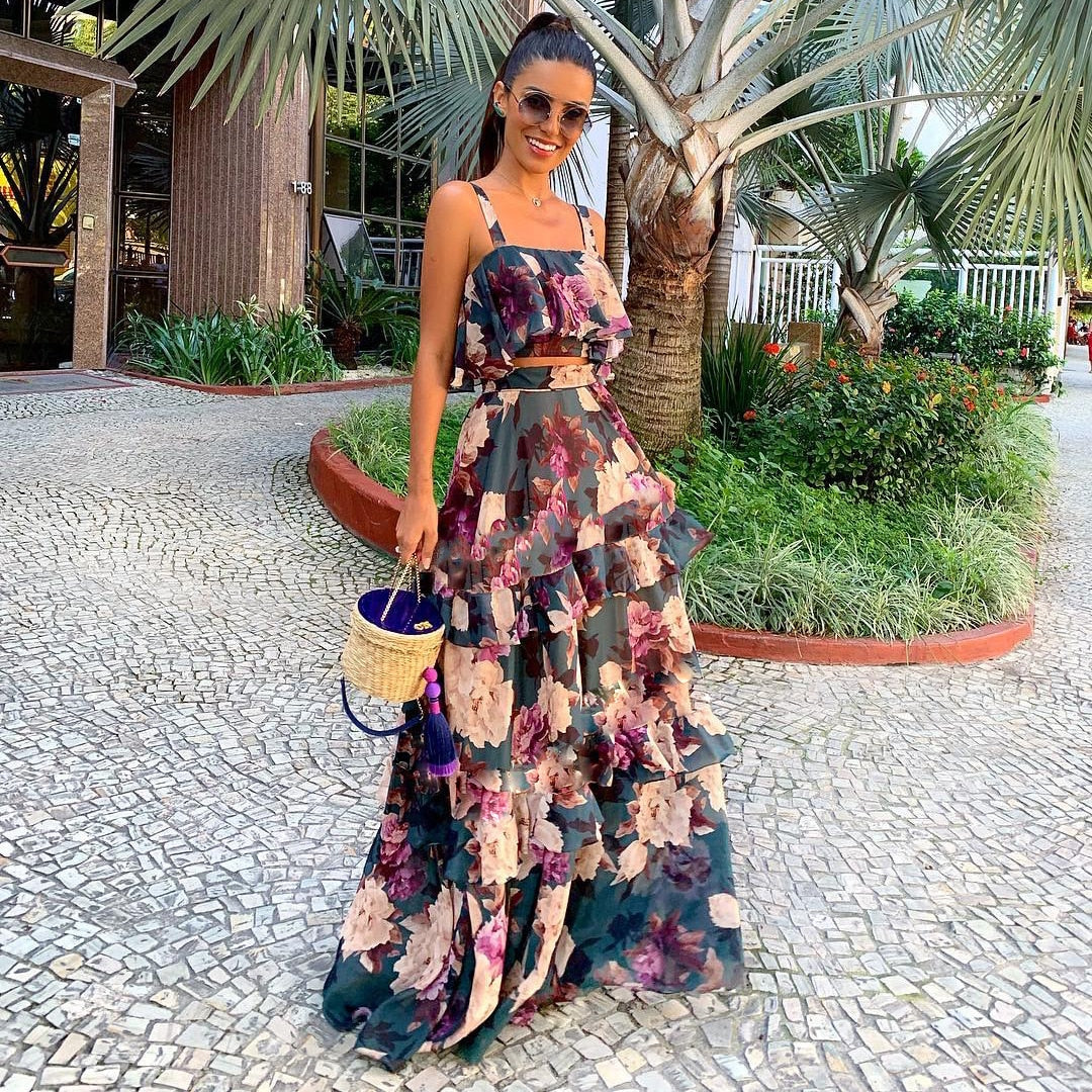 Frau in langem, geblümtem Sommerkleid mit Korbtasche, Sonnenbrille, draußen, tropische Umgebung.
