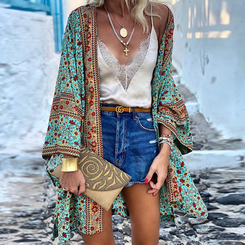 Frau in Boho-Kimono, blau-rot gemustert, mit Jeansshorts und goldener Clutch auf gepflastertem Weg.