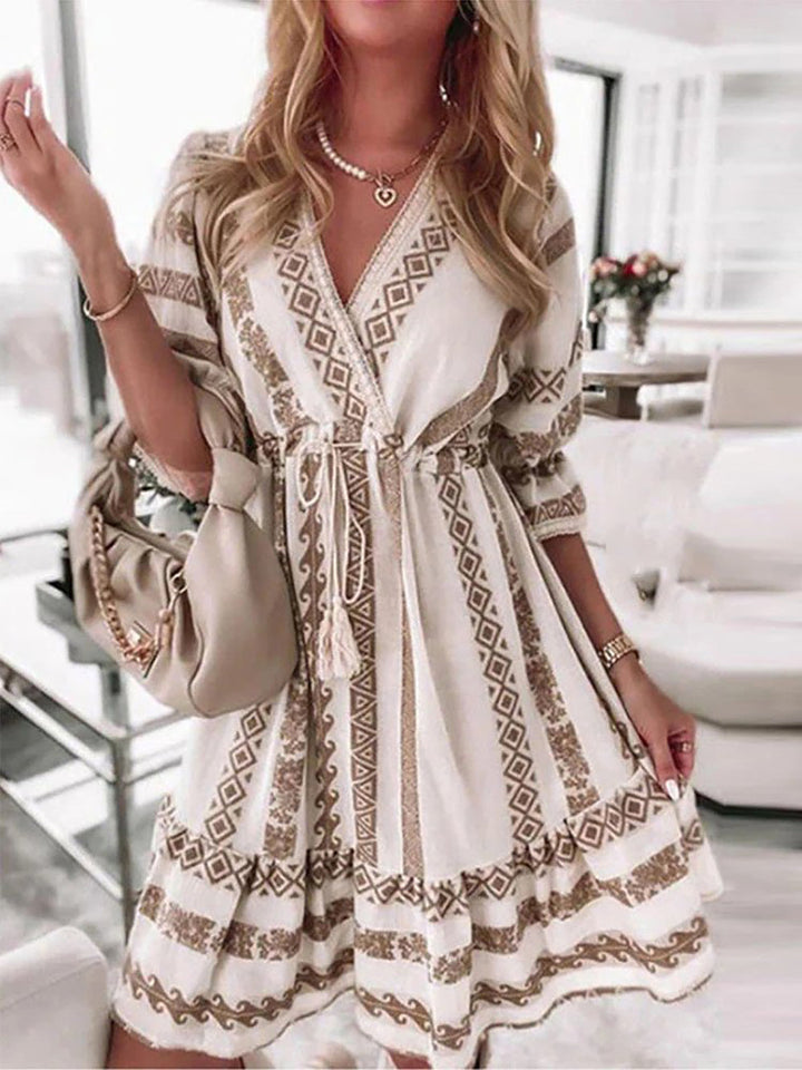 Frau in weißem Boho-Kleid mit geometrischem Muster, beige Handtasche, elegantes Wohnzimmer.