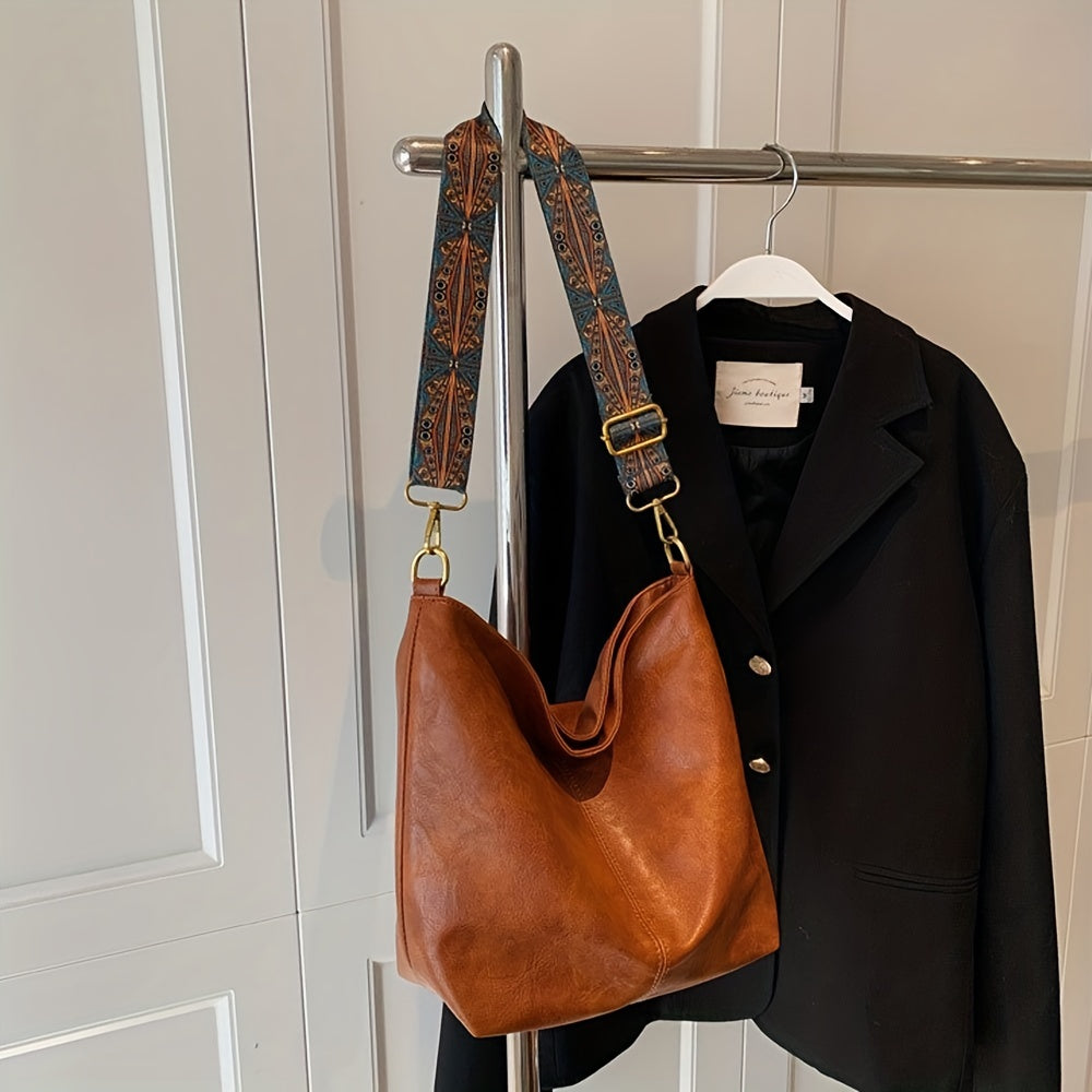 Braune Ledertasche mit gemustertem Schultergurt, hängt an schwarzem Blazer auf Kleiderstange.