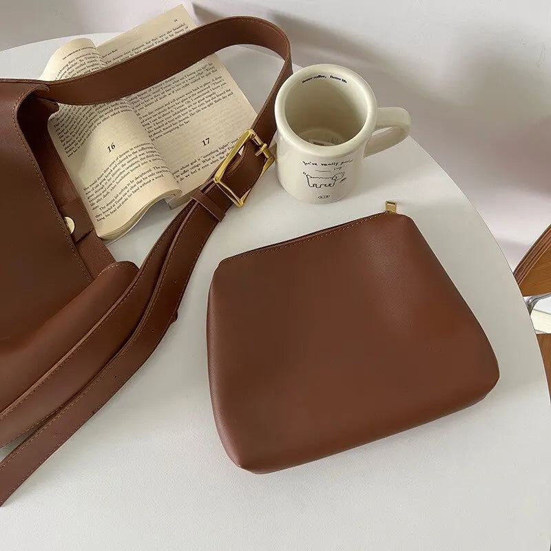 Braune Ledertasche mit passender Geldbörse auf weißem Tisch, Kaffeetasse und Buch im Hintergrund.