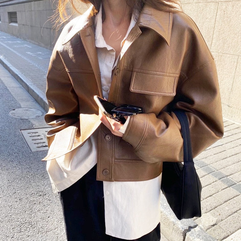 Frau in brauner Lederjacke, weißem Hemd, schwarze Tasche, Sonnenbrille, Straße, modisch.