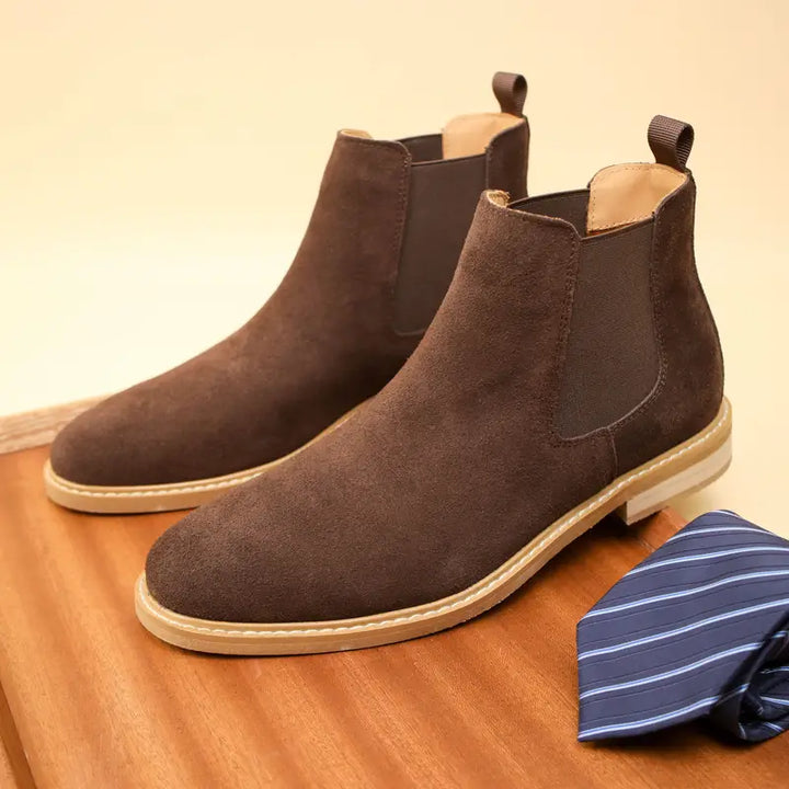 Braune Wildleder-Chelsea-Boots auf Holztisch neben gestreifter Krawatte.