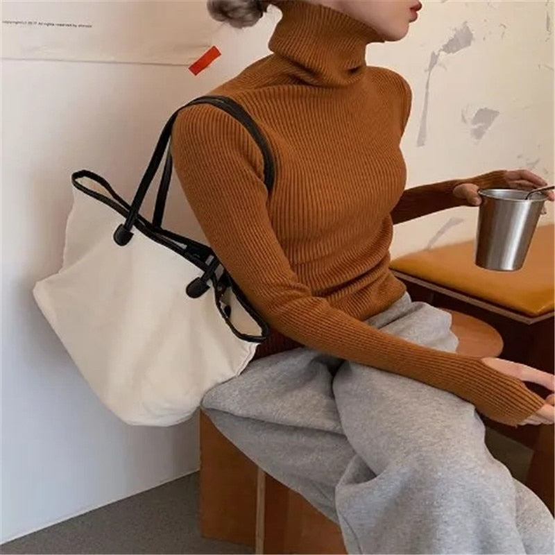 Frau in braunem Rollkragenpullover mit weißer Stofftasche und grauer Hose, hält Metallbecher.