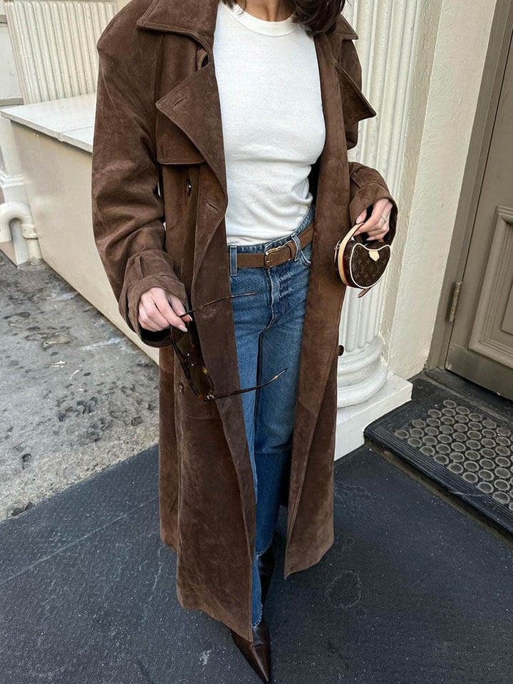 Frau in braunem Wildledermantel, weißem Shirt, blauer Jeans, hält Sonnenbrille und Herz-Tasche.