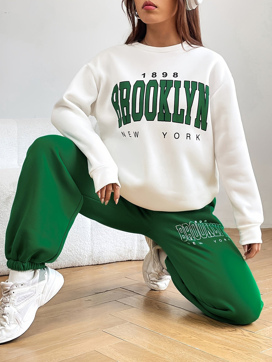 Frau in weißem Brooklyn-Pullover und grüner Jogginghose, kniend auf Holzboden.