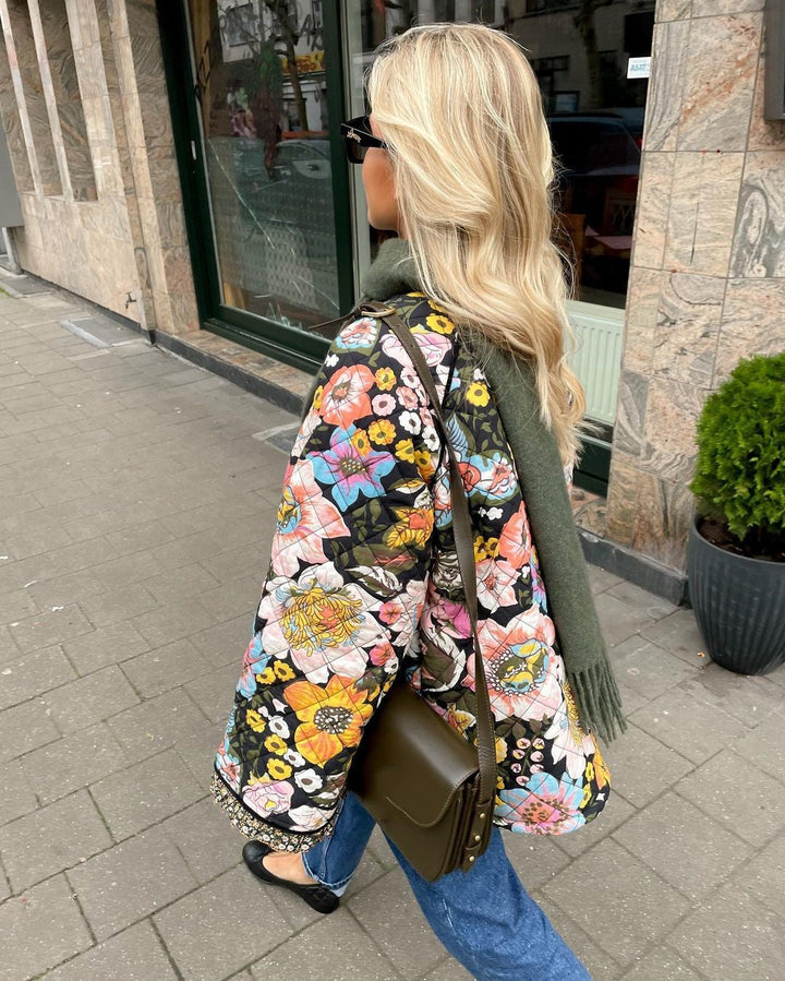 Frau mit bunter Blumenjacke, grüner Tasche und Schal spaziert auf Bürgersteig.