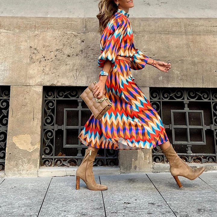 Frau in bunter Midi-Kleid mit geometrischem Muster, braune Stiefel, beige Tasche, Stadtstraße.