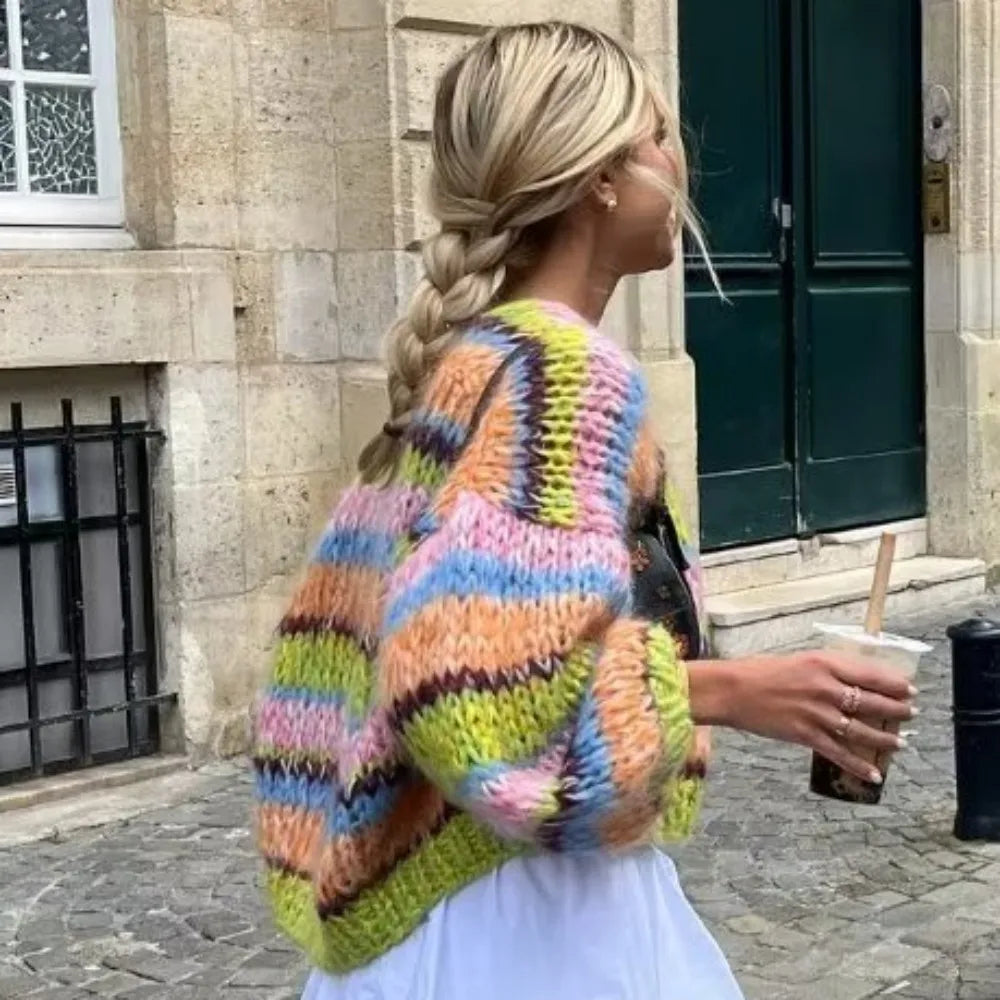 Frau mit buntem Strickpullover, Zopf, hält Getränk, läuft auf Kopfsteinpflaster vor Gebäude.
