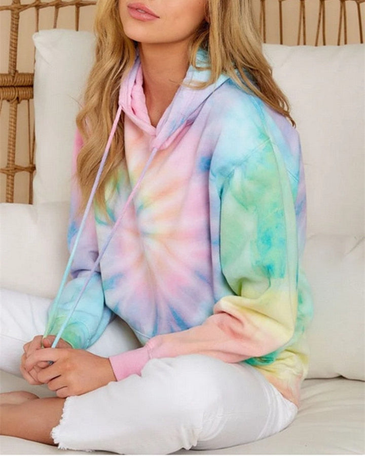 Frau in pastellfarbenem Batik-Hoodie, sitzt auf weißem Sofa, lässige Mode, gemütlicher Stil.
