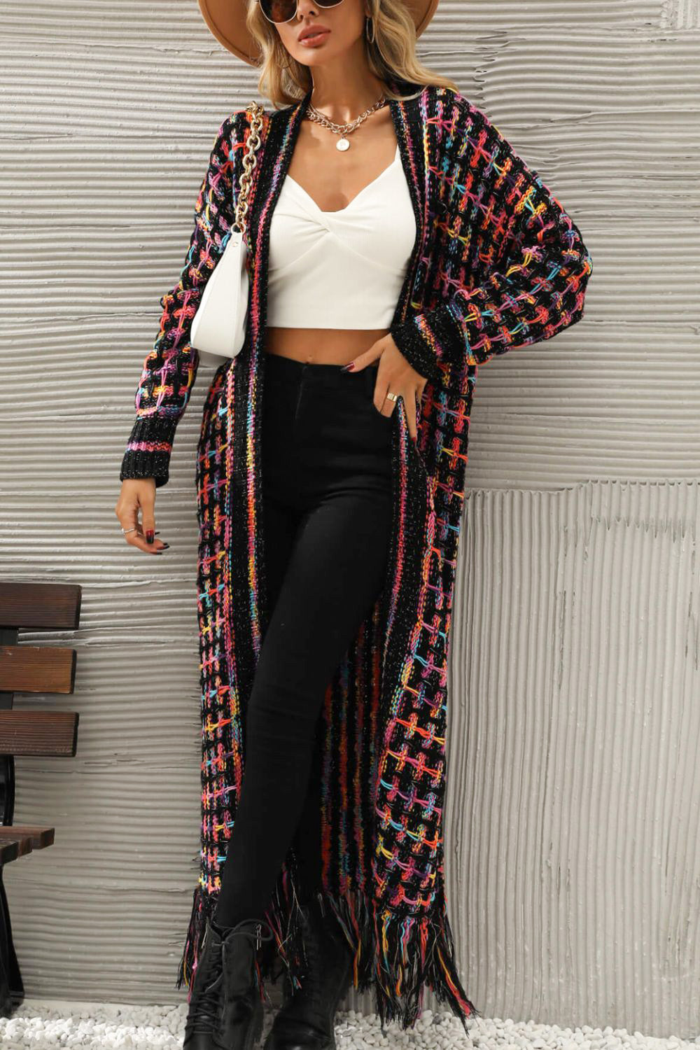 Frau in langem, buntem Strickmantel, weißem Top, schwarzer Hose, Hut und Sonnenbrille.