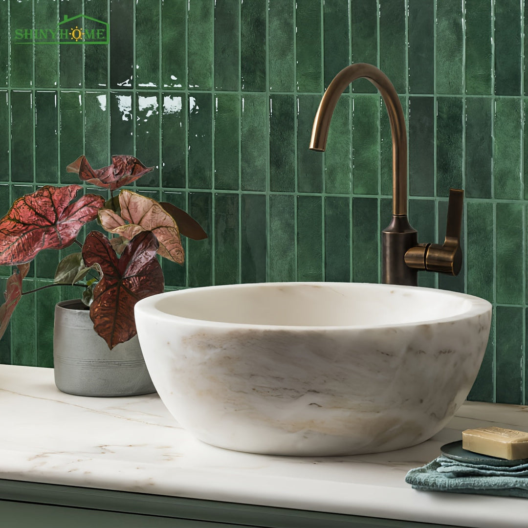 Weiße Marmor-Waschschüssel mit bronzefarbenem Wasserhahn, grüne Fliesen, Zimmerpflanze.