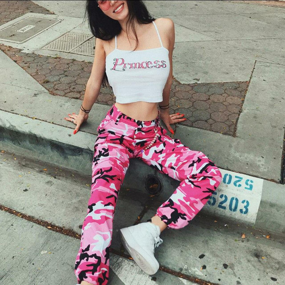 Frau in rosa Camouflage-Hose und weißem "Princess"-Top, sitzt auf Bürgersteig.