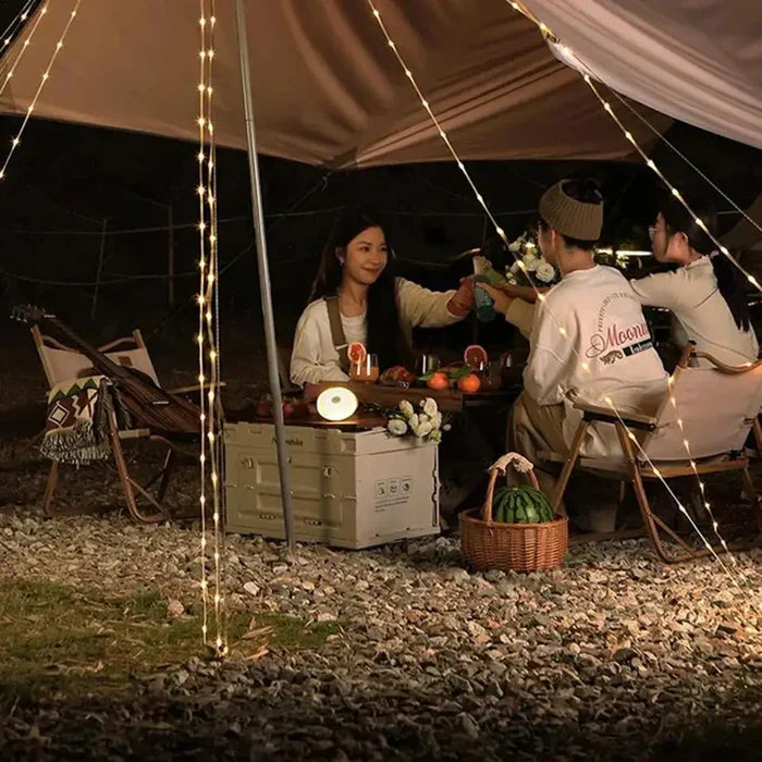 Gruppe beim Camping unter Lichterkette, sitzt um Tisch mit Essen und Getränken, Abendstimmung.