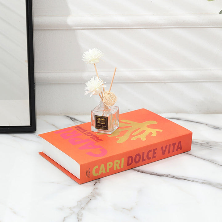 Oranges Buch "Capri Dolce Vita" auf Marmortisch mit Duftflakon und weißen Blumen.
