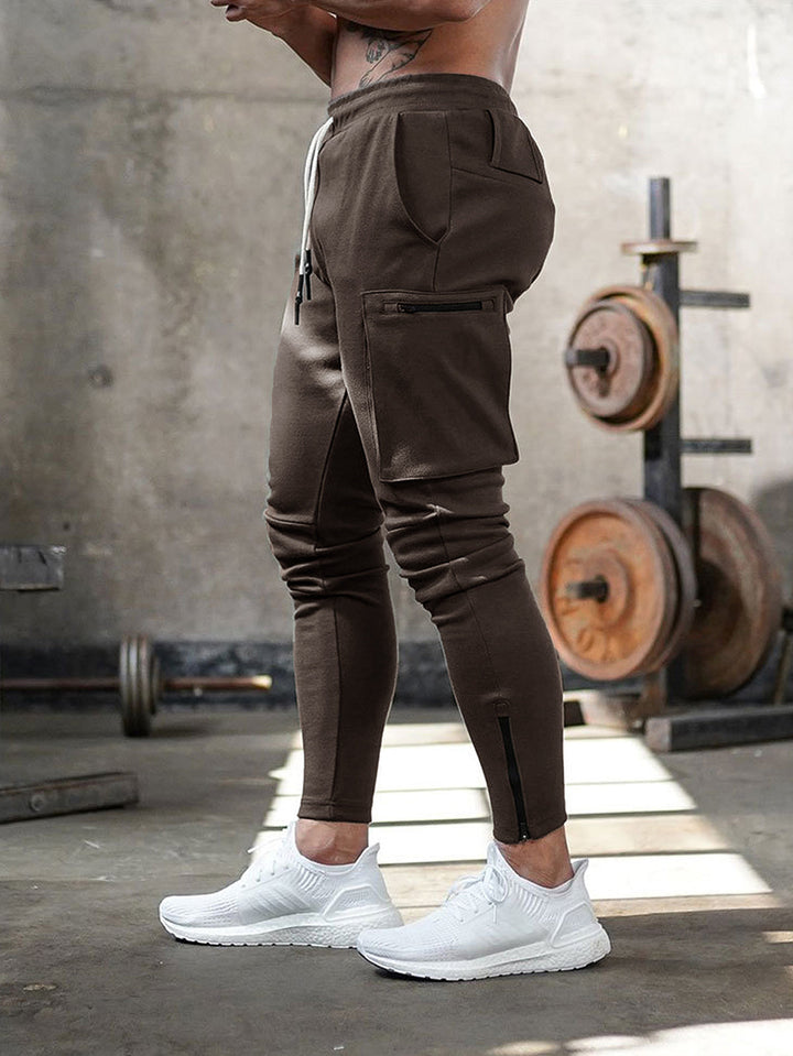 Mann in brauner Cargo-Jogginghose im Fitnessstudio, weiße Sneaker, Hantel im Hintergrund.