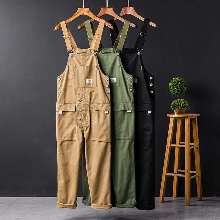 Drei Herren-Latzhosen in Beige, Grün und Schwarz auf Kleiderbügeln vor schwarzem Hintergrund.