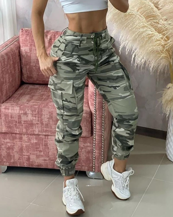 Frau in Camouflage-Cargohose und weißem Top, steht neben rosa Sofa.