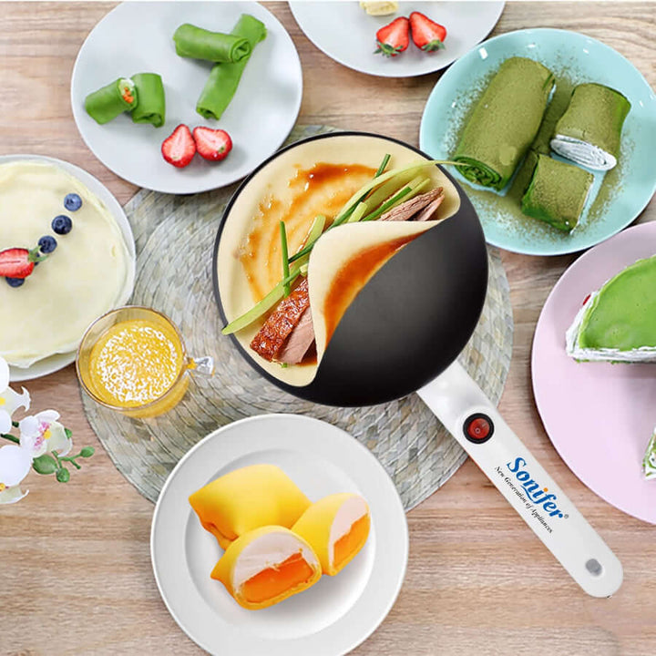 Elektrische Crêpe-Maker mit weißem Griff, umgeben von gefüllten Crêpes und Obst.