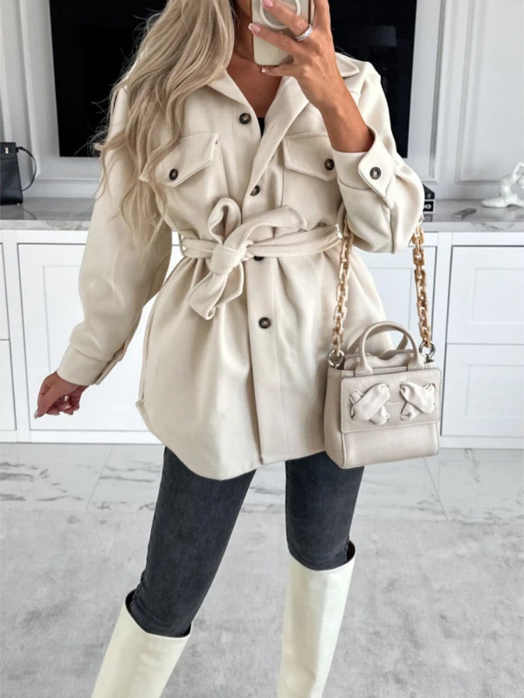 Frau in cremefarbener Jacke mit Gürtel, grauer Hose, weißer Stiefel, beige Handtasche.